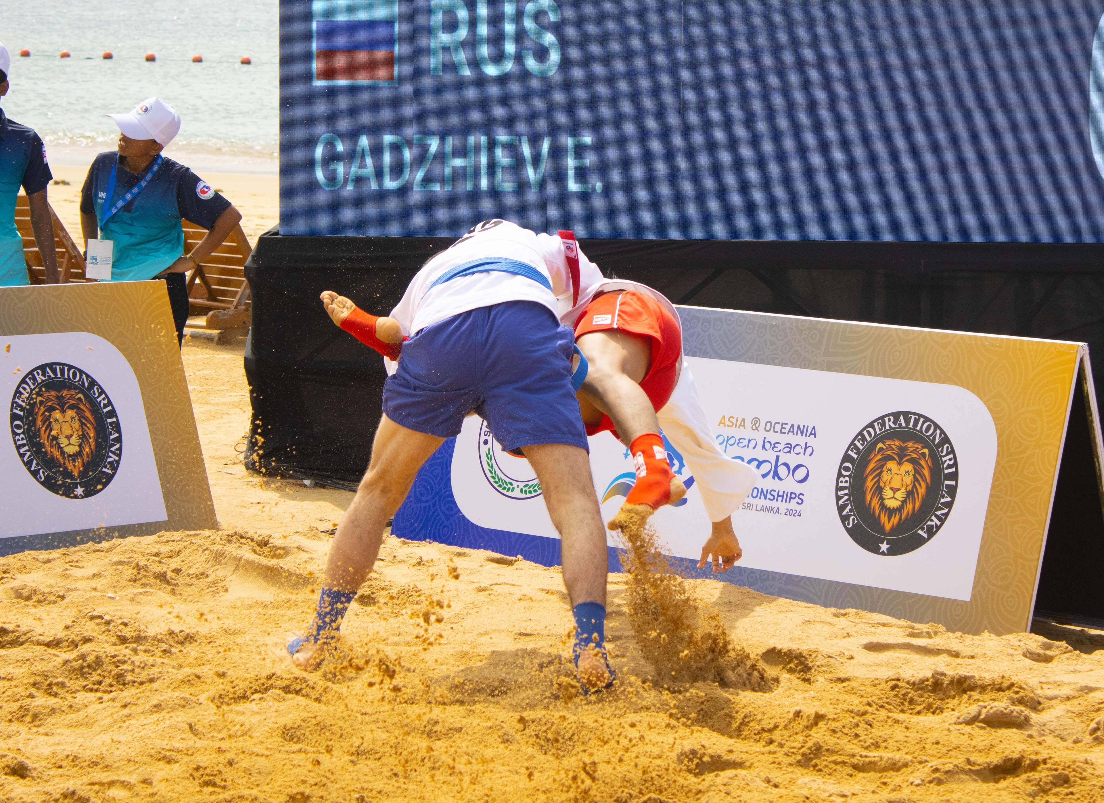 A throw mid-execution, sand spraying across the pit as the scoreboard reads out the bout